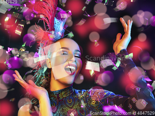 Image of Beautiful young woman in carnival and masquerade costume in colorful neon lights on black background in flying confetti