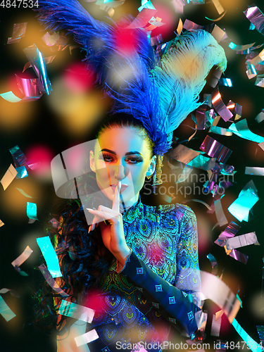 Image of Beautiful young woman in carnival and masquerade costume in colorful neon lights on black background in flying confetti