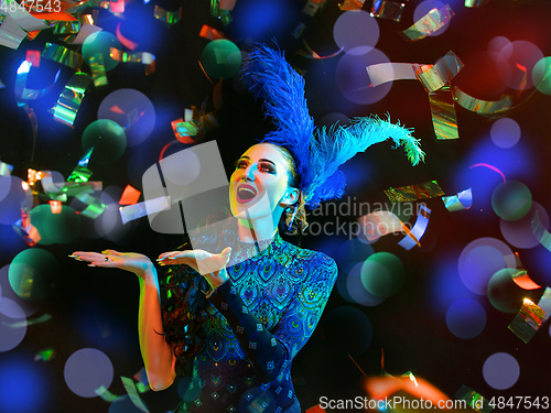 Image of Beautiful young woman in carnival and masquerade costume in colorful neon lights on black background in flying confetti