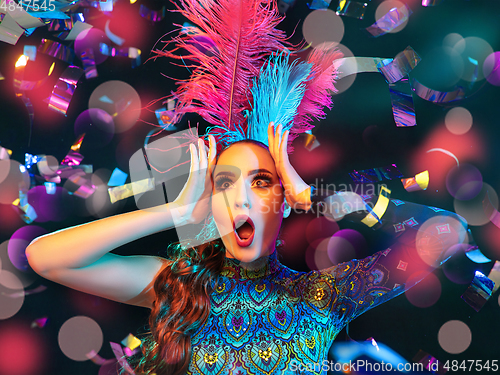 Image of Beautiful young woman in carnival and masquerade costume in colorful neon lights on black background in flying confetti