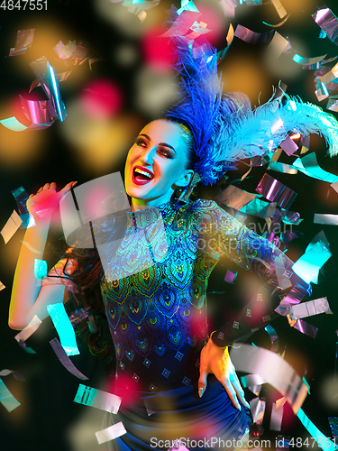 Image of Beautiful young woman in carnival and masquerade costume in colorful neon lights on black background in flying confetti