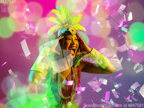 Image of Beautiful young woman in carnival and masquerade costume in colorful neon lights on gradient background in flying confetti