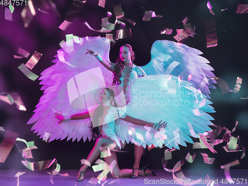 Image of Young female dancers with angel\'s wings in neon light on black background in flying confetti