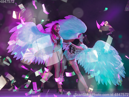 Image of Young female dancers with angel\'s wings in neon light on black background in flying confetti