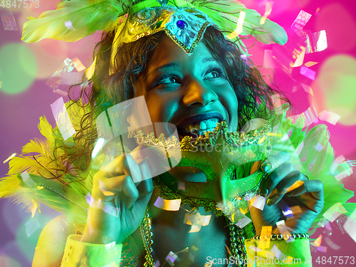 Image of Beautiful young woman in carnival and masquerade costume in colorful neon lights on gradient background in flying confetti