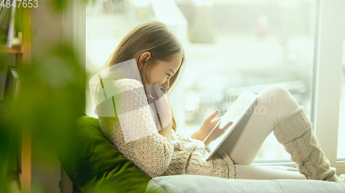 Image of Little girl sitting near by window with tablet, remote studying, being at home