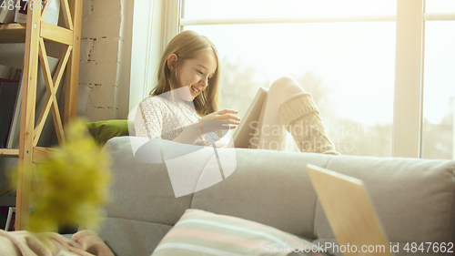 Image of Little girl sitting near by window with tablet, remote studying, being at home