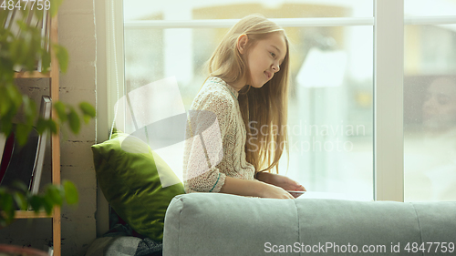Image of Little girl sitting near by window with tablet, remote studying, being at home