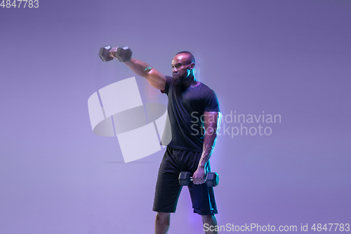 Image of Young african-american bodybuilder training over purple background in neon, mixed light