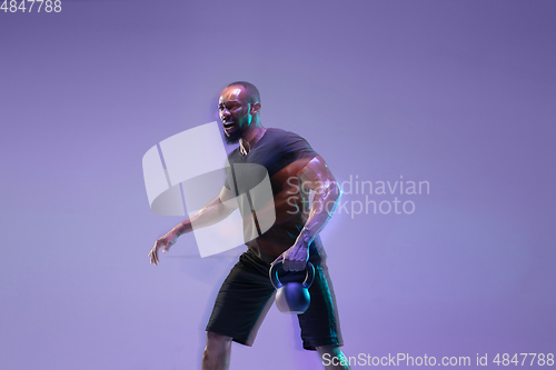 Image of Young african-american bodybuilder training over purple background in neon, mixed light