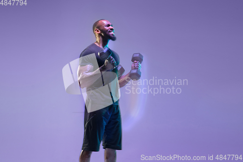 Image of Young african-american bodybuilder training over purple background in neon, mixed light