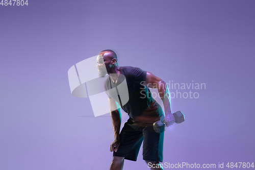Image of Young african-american bodybuilder training over purple background in neon, mixed light