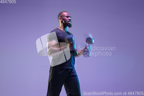 Image of Young african-american bodybuilder training over purple background in neon, mixed light