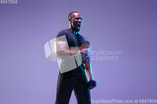 Image of Young african-american bodybuilder training over purple background in neon, mixed light