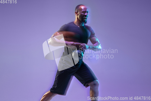 Image of Young african-american bodybuilder training over purple background in neon, mixed light