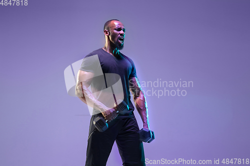 Image of Young african-american bodybuilder training over purple background in neon, mixed light