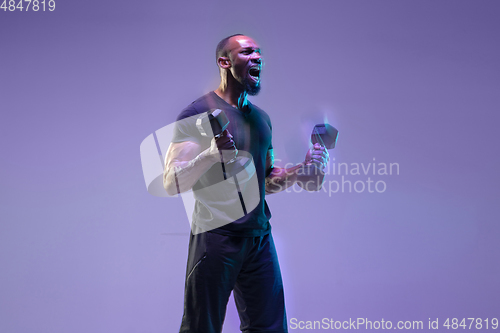 Image of Young african-american bodybuilder training over purple background in neon, mixed light