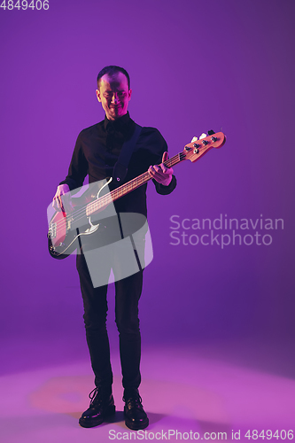 Image of Young caucasian musician playing guitar in neon light on purple background