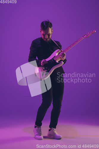Image of Young caucasian musician playing guitar in neon light on purple background