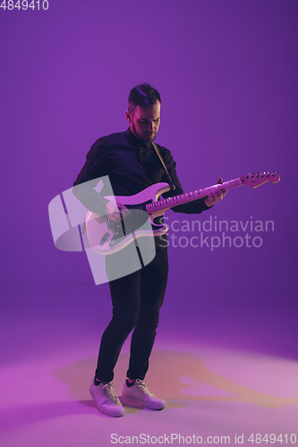 Image of Young caucasian musician playing guitar in neon light on purple background