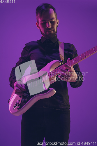 Image of Young caucasian musician playing guitar in neon light on purple background