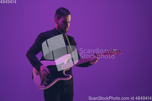 Image of Young caucasian musician playing guitar in neon light on purple background