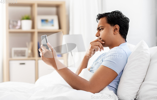 Image of thinking indian man with smartphone in bed at home