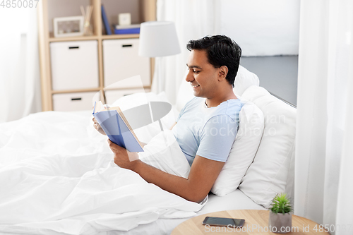 Image of happy indian man reading book in bed at home