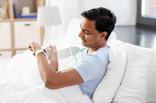 Image of happy indian man with smart watch in bed at home