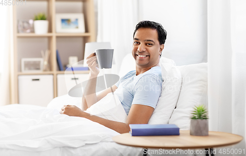 Image of happy indian man drinking coffee in bed at home