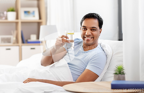 Image of happy indian man drinking water lying in bed