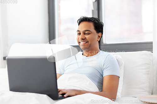 Image of happy indian man with laptop in bed at home