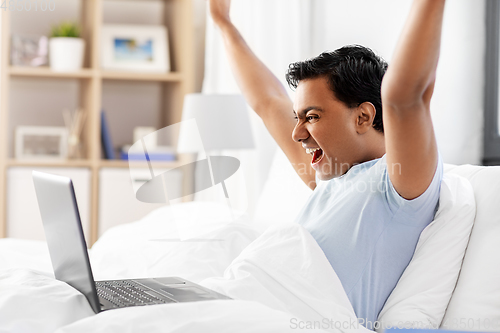 Image of happy indian man with laptop in bed at home