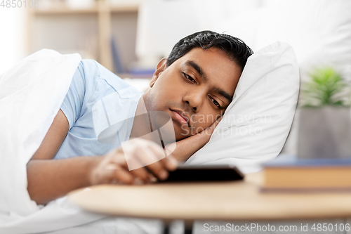 Image of sleepy indian man in bed looking at smartphone