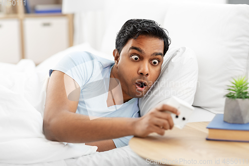 Image of overslept indian man in bed looking at alarm clock