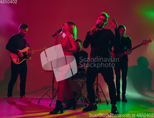 Image of Young caucasian musicians, band performing in neon light on red studio background