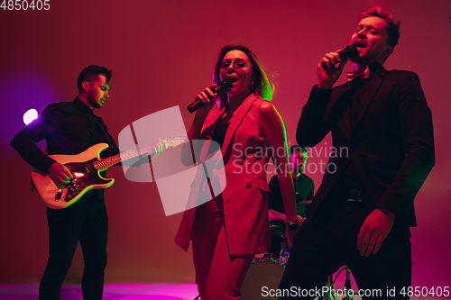 Image of Young caucasian musicians, band performing in neon light on red studio background