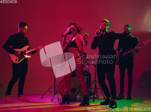 Image of Young caucasian musicians, band performing in neon light on red studio background