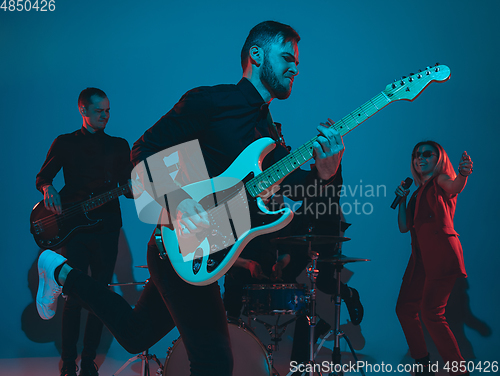 Image of Young caucasian musicians, band performing in neon light on blue studio background, guitarist in front