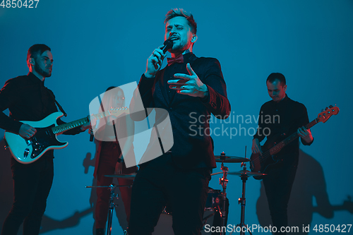 Image of Young caucasian musicians, band performing in neon light on blue studio background, singer in front