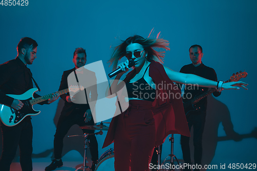 Image of Young caucasian musicians, band performing in neon light on blue studio background, singer in front