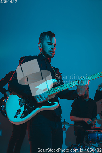 Image of Young caucasian musicians, band performing in neon light on blue studio background, guitarist in front