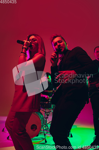 Image of Young caucasian musicians, singers performing in neon light on red studio background