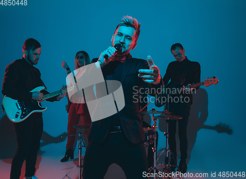 Image of Young caucasian musicians, band performing in neon light on blue studio background, singer in front
