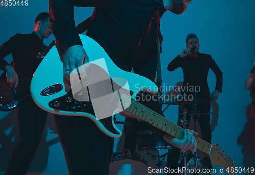 Image of Young caucasian musicians, band performing in neon light on blue studio background, guitarist in front