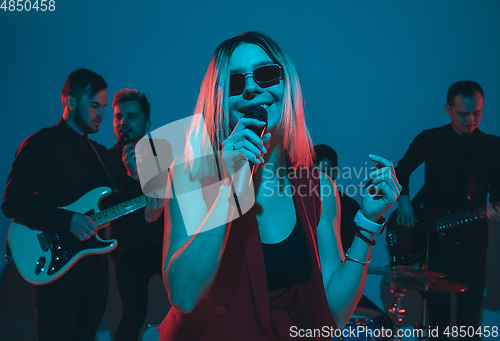 Image of Young caucasian musicians, band performing in neon light on blue studio background, singer in front