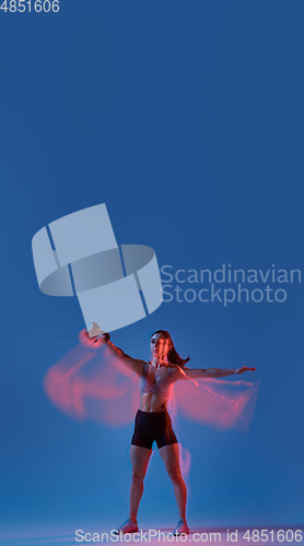 Image of Caucasian professional female athlete training on blue studio background in neon, mixed light. Muscular, sportive woman.