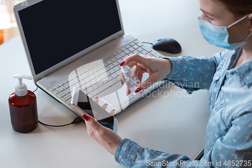 Image of Young woman in face mask disinfecting gadgets surfaces on her workplace