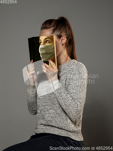 Image of Be safe and read to become someone else - woman covering face with book in face mask while reading on grey background