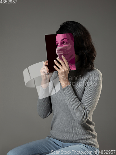 Image of Be safe and read to become someone else - woman covering face with book in face mask while reading on grey background